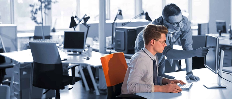 A man is working on a computer in an office. Standing next to him is an experienced IT advisor pointing at the screen to show the benefits they have achieved.