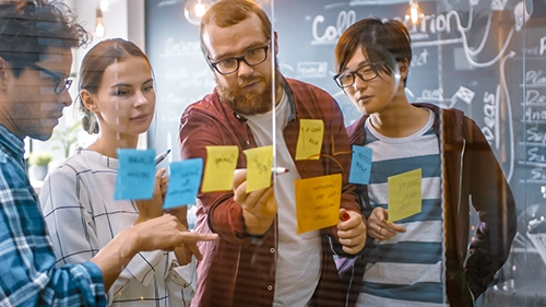 A group of people looking at sticky notes on a wall while they try to solve a complex problem.